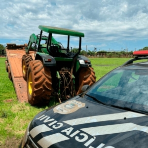 Viatura da Polícia Civil em primeiro plano e trator agrícola sobre caminhão ao fundo, ilustrando uma operação de busca e apreensão de maquinário.